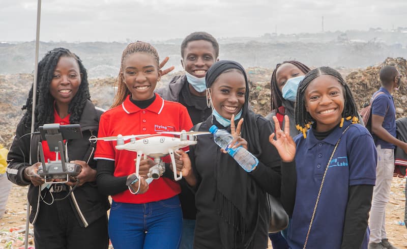 five girls are holding drones in front of a rocky beach. Two of them and a boy behind them hold up peace signs.