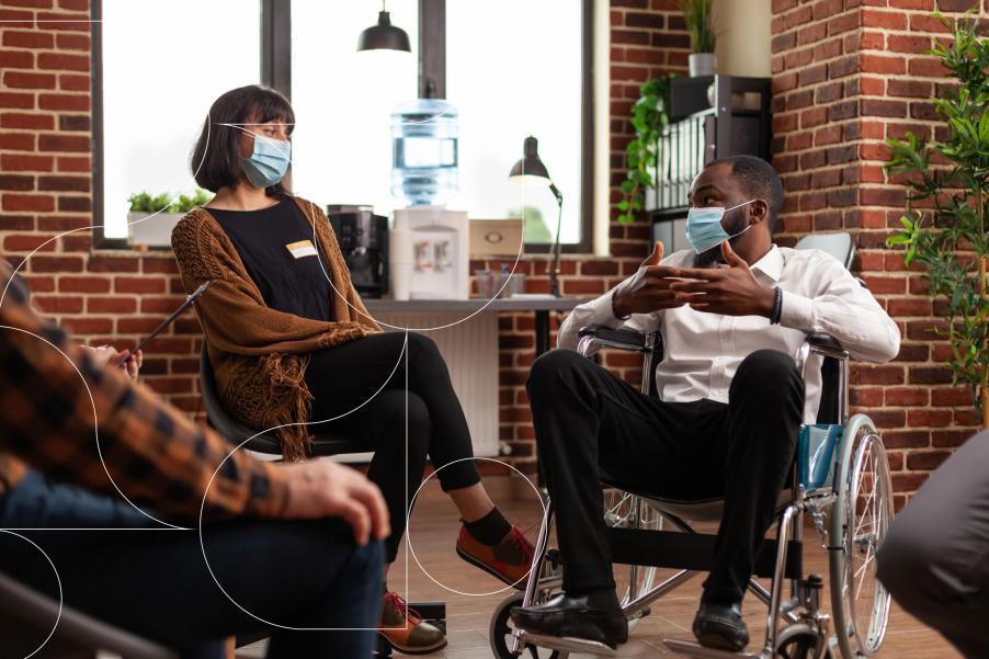 A group of people sitting in a circle. Only two of individuals are fully in frame, a woman with short black hair and a black man in a wheelchair, both wearing medical masks.