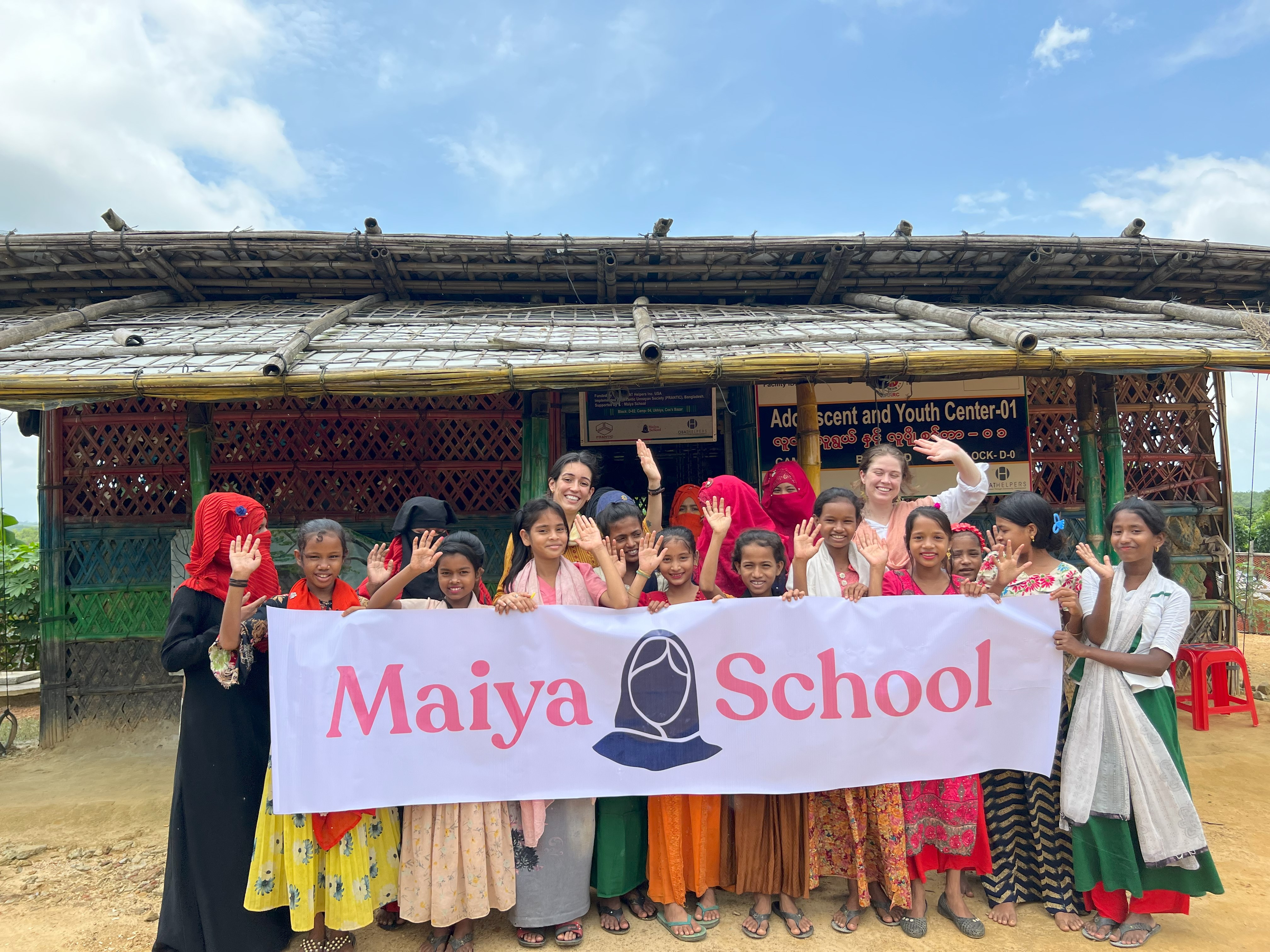 A group of girls stands in front of a building. They are holding a banner that reads "Maiya School"