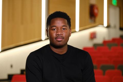 A African black man wearing a black shirt poses sitting in an auditorium. The photo is taken in a portrait style.