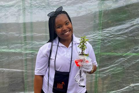 A young black woman stands smiling in front of a greenhouse. She is wearing a white shirt and white gardening gloves holding a small jade plant in one hand.