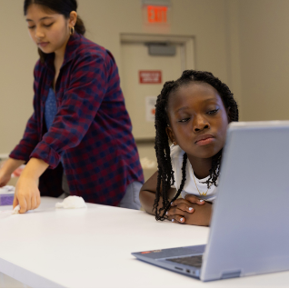 A young black girl studies the screen of a laptop while a young female educator organizes lab materials behind her.