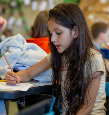 A young girl with long brown hair is writing on a piece of paper at her desk. In the background, out of focus, other students seem to be doing the same.