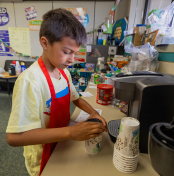 A young boy in a red apron is at his classroms snack station, carefully preparing a hot beverage in a disposable cup.